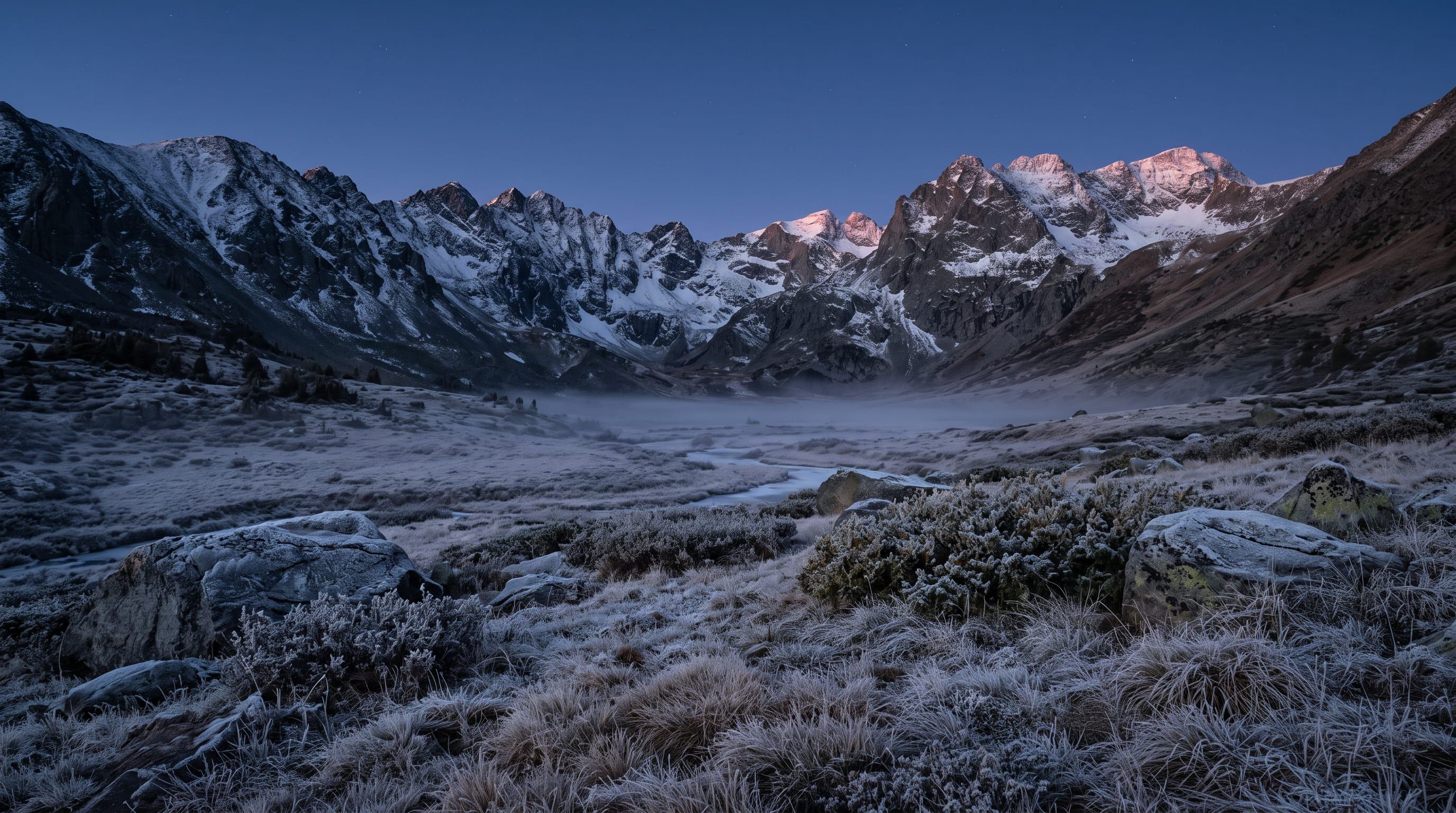 Een stille hoogalpiene kom in het koude blauwe licht vlak vóór zonsopkomst, met rijp op het gras en besneeuwde bergkammen die de eerste warme gloed vangen.