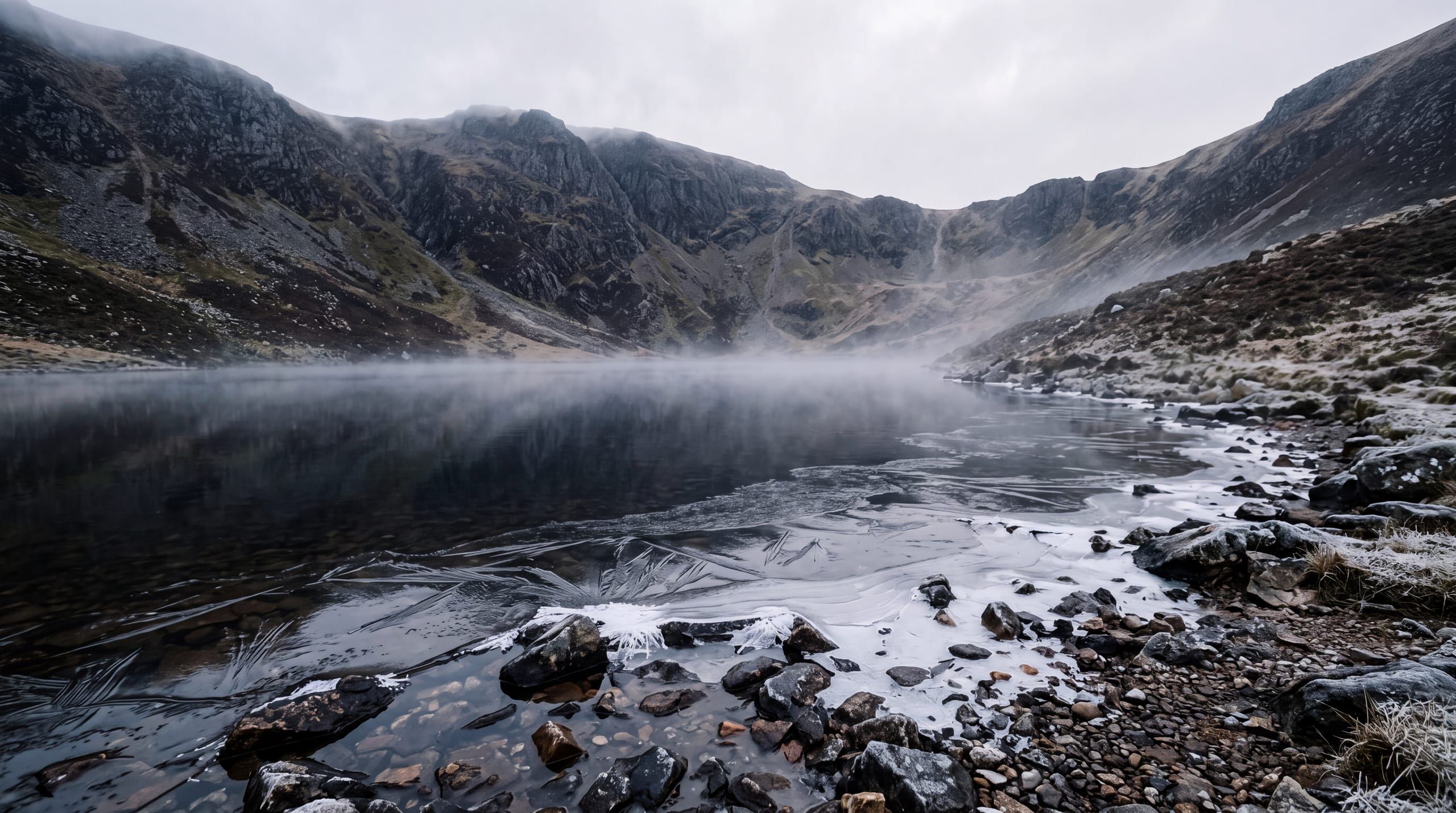 Een bergmeertje dat bij dageraad dichtvriest: dunne ijsplaten en veervormige kristallen over stil donker water, met mist op het oppervlak en de bergkom erachter in de wolken.