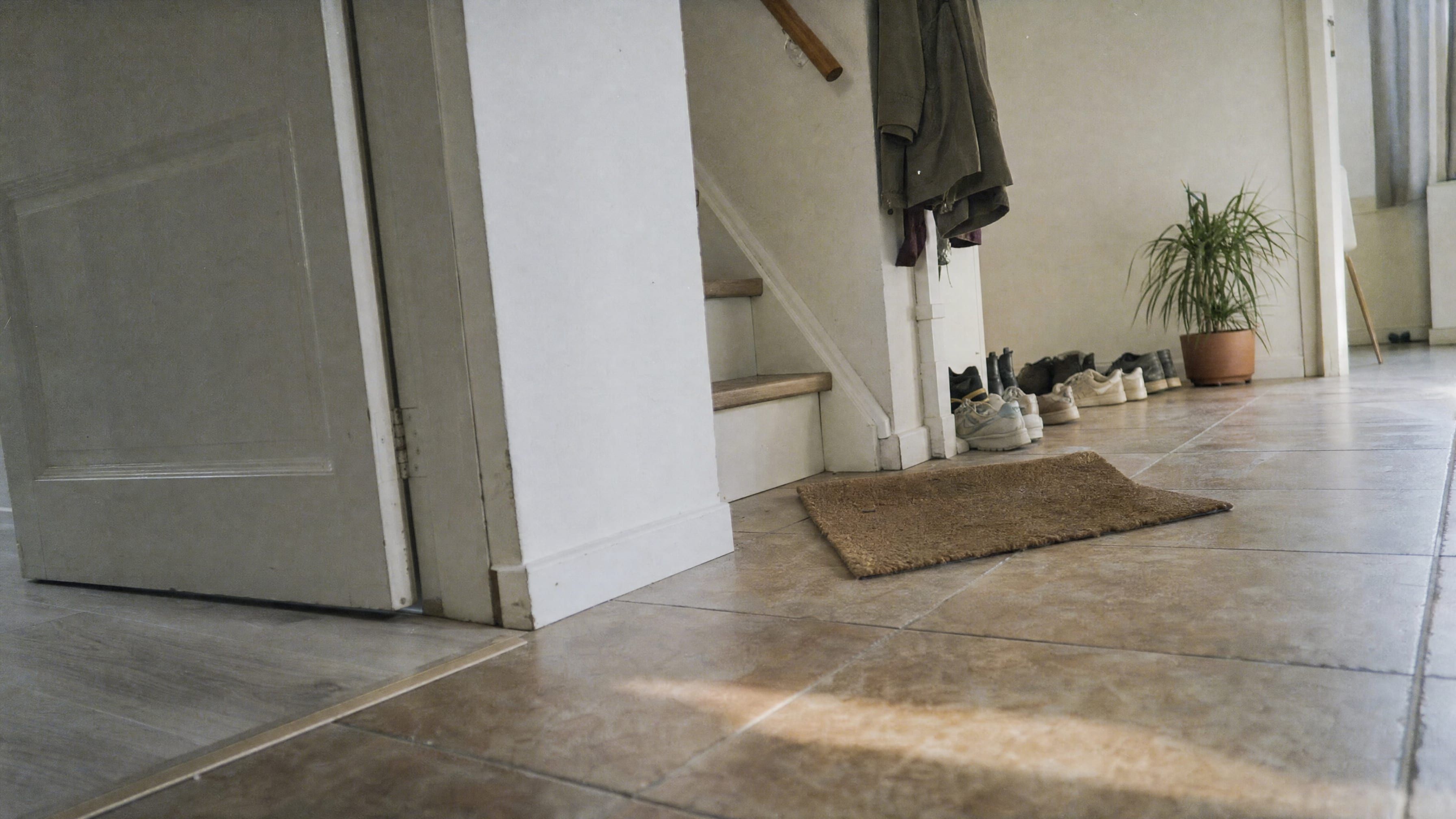 Een gang in een gewone woning, laag bij de vloer gezien: een tegelvloer met een smalle drempelstrip in de deuropening, een kokosmat, onderaan de trap enkele paren schoenen op een rij en jassen aan de wand.